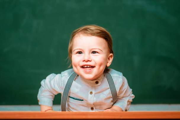 Friendly child in classroom near blackboard desk. Little student. Cute preschooler is sitting at a desk indoors. Individual tutoring - Foto, Bild