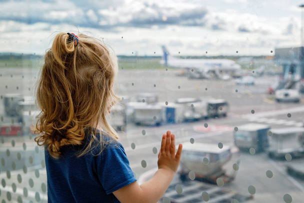 Cute little toddler girl at the airport, traveling. Happy healthy child waiting near window and watching airplanes. Family going on summer vacations by plane - Zdjęcie, obraz