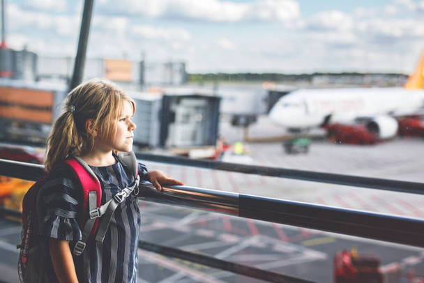 Little girl at the airport is waiting for landing at the big window. Cute kid stands at the window against the backdrop of airplanes. Looking forward to leaving for a family summer vacation - Photo, Image