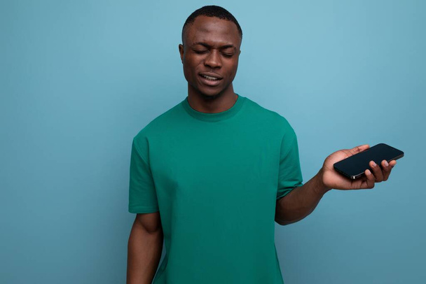 Tişört giyen yakışıklı Afrikalı genç adam sosyal medya için akıllı telefonu fotokopi alanı ile izole edilmiş arka planda kullanıyor.. - Fotoğraf, Görsel