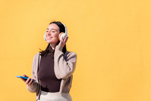 woman enjoying happy listening to music from mobile phone in headphones with a yellow wall in the background, concept of rhythm and positive people, copy space for text - Фото, изображение