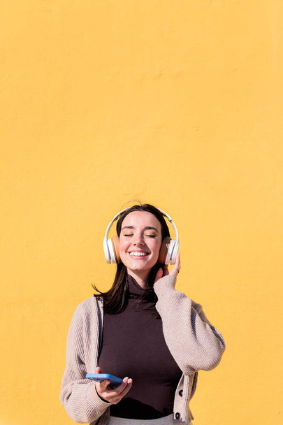 woman enjoying happy listening to music from mobile phone in headphones with a yellow wall in the background, concept of rhythm and positive people, copy space for text - Фото, изображение
