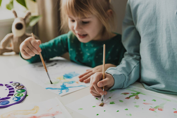 Two cute little sisters or classmates are painting on table with brush. Small girls are laughing. Kids are in kindergarten or at home. Child in living room. Home schooling concept, doing homework  - Photo, Image