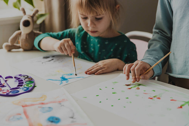 Two cute little sisters, classmates are painting on table. Small preschool and school girl. Kids use brush in kindergarten or school. Adorable preschooler in living room. Home schooling concep - Photo, Image