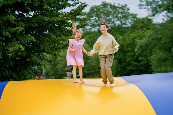 Little preschool girl and school sister jumping on trampoline. Happy funny children, siblings in love having fun with outdoor activity in summer. Trampolin in ukrainian flagg colors. - Photo, Image