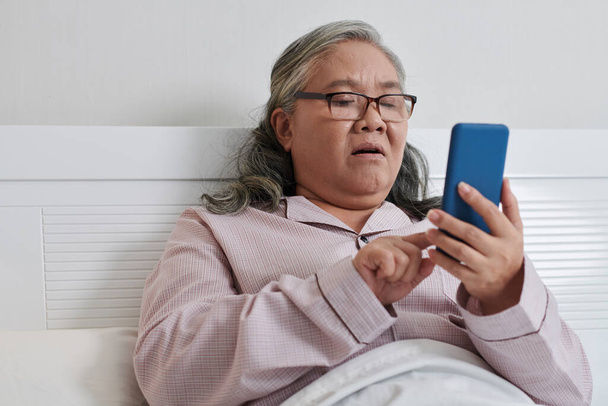 Femme âgée choquée assise au lit et lisant les nouvelles sur smartphone - Photo, image