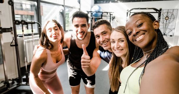 Group of diverse multiethnic young friends in sportswear smiling and showing thumbs up while hugging each other and taking self portrait in sports club - Photo, Image