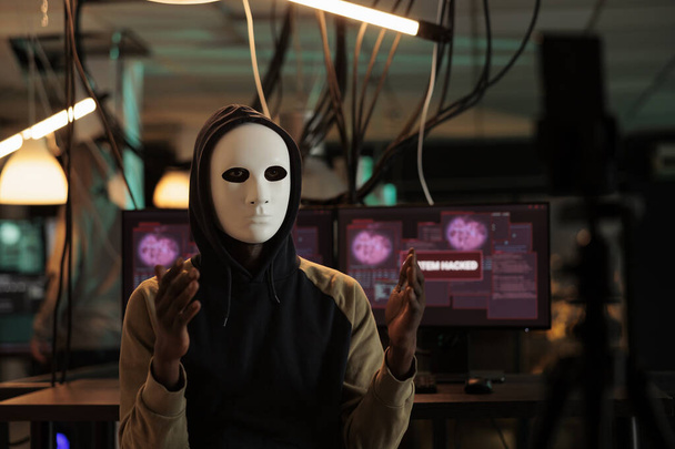 African american hacker broadcasting threat video at night, wearing anonymous mask and asking ransomware. Male spy stealing database information with trojan virus and computer malware. - Foto, Imagem