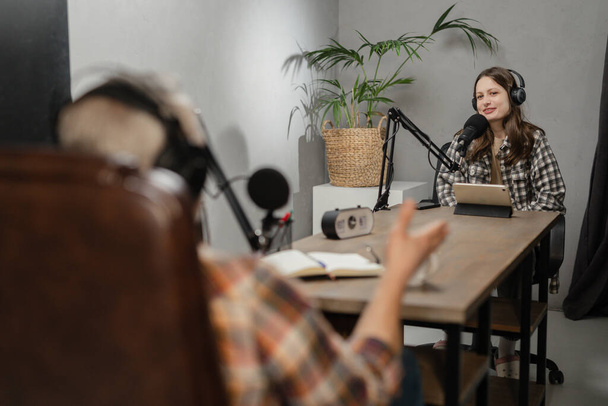Een jonge vrouw met een koptelefoon zit aan een tafel voor een microfoon en organiseert een gezamenlijke podcast met een uitgenodigde gast. Medewerkers van het entertainment radiostation zijn samen in de ether. Hoge kwaliteit - Foto, afbeelding