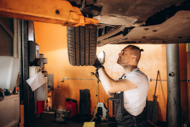Механик осматривает шины автомобиля с помощью фонарика в ремонтном гараже - Фото, изображение