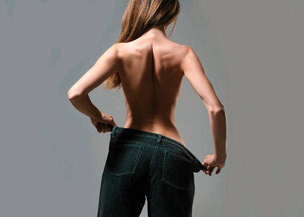 Slim girl have points at slim waist in big trousers, successful weight loss, isolated on gray background. Successful weight loss, woman with too large jeans after a diet. Skinny waist, back view - Foto, Bild