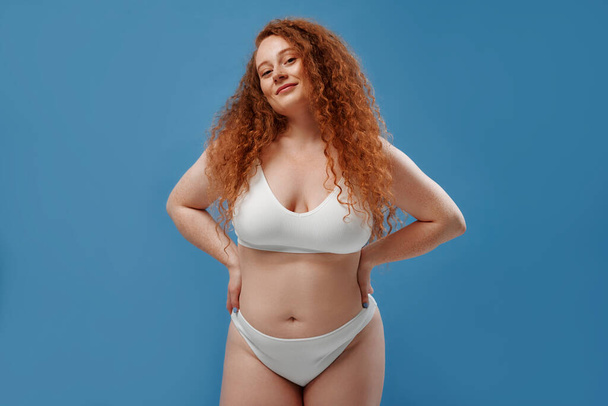 Young pretty redhead plus size or plump woman celebrating her natural body. Positive beautiful female model in white underwear against blue background. Lifestyle portrait with minimal makeup - Valokuva, kuva
