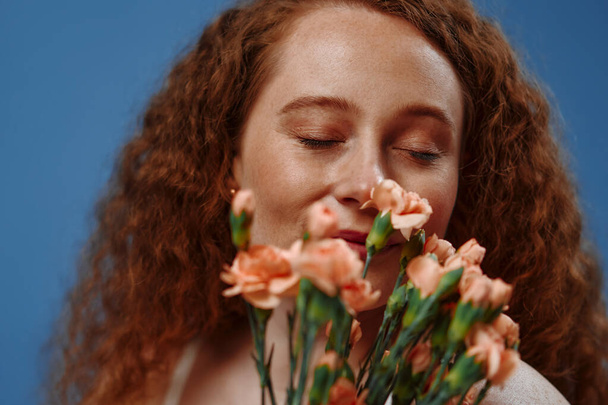 Young redhead plus size or plump woman with flowers celebrating her natural body. Positive beautiful female model in white underwear against blue background. Cosmetology, skin and body care concept. - Foto, afbeelding