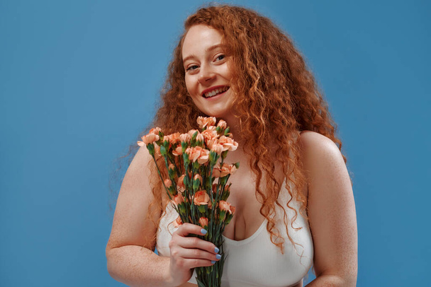 Young redhead plus size or plump woman with flowers celebrating her natural body. Positive beautiful female model in white underwear against blue background. Cosmetology, skin and body care concept. - 写真・画像