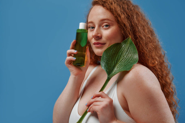 Beautiful redhead plus size woman with freckles in white lingerie with bottle of lotion and leaf isolated on blue background. Body positive. Cosmetology and skin care. - Foto, Imagem