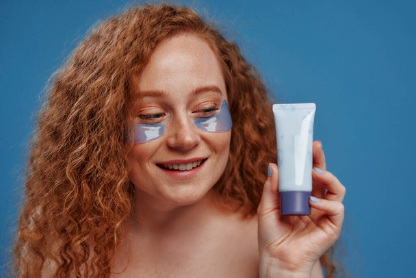 Beautiful redhead plus size woman with freckles using eye patches and holding tube of cream isolated on blue background. Body positive. Cosmetology and skin care. - Foto, Imagem