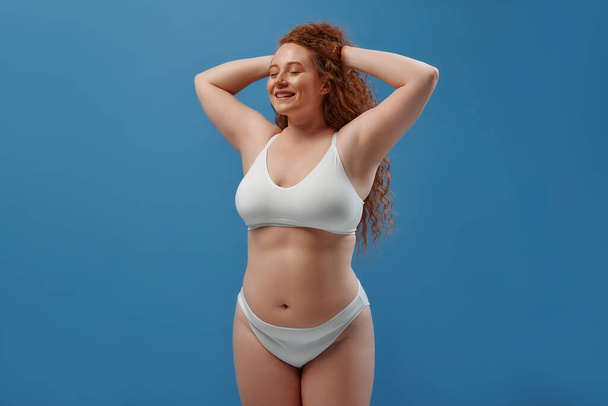 Young pretty redhead plus size or plump woman celebrating her natural body. Positive beautiful female model in white underwear against blue background. Lifestyle portrait with minimal makeup - Fotoğraf, Görsel