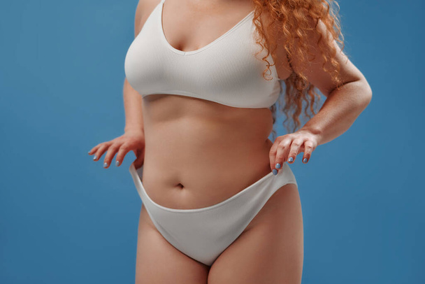 Young pretty redhead plus size or plump woman celebrating her natural body. Positive beautiful female model in white underwear against blue background. Lifestyle portrait with minimal makeup - Photo, image