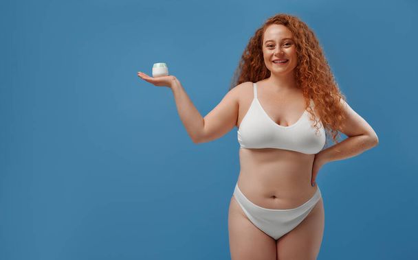 Redhead freckled young plump plus size woman in white lingerie holding palm with jar of moisturizer. Body positive, skin care and cosmetology. - Φωτογραφία, εικόνα