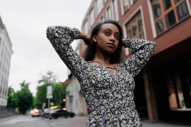 La mujer joven africana pensativa en el vestido veraniego pasea en la ciudad sobre el fondo del edificio. Vista inferior. - Foto, Imagen