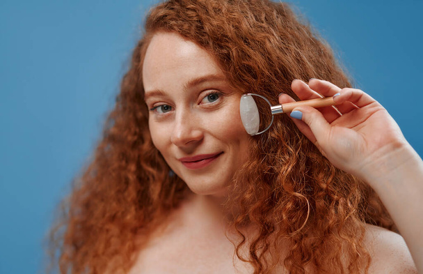 Freckled redhead plump plus size woman in white lingerie using face roller for facelift isolated on blue background. The concept of skin care, body positive and cosmetology. - Fotografie, Obrázek