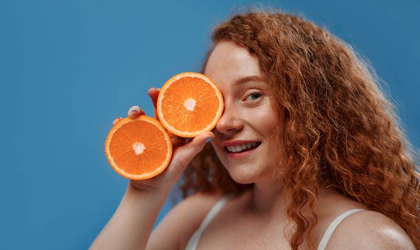 Piegowata ruda pulchna plus size kobieta w białej bieliźnie trzymająca pomarańczowe połówki odizolowane na niebieskim tle. Koncepcja pielęgnacji twarzy i ciała, ciała pozytywnego i kosmetologii. - Zdjęcie, obraz