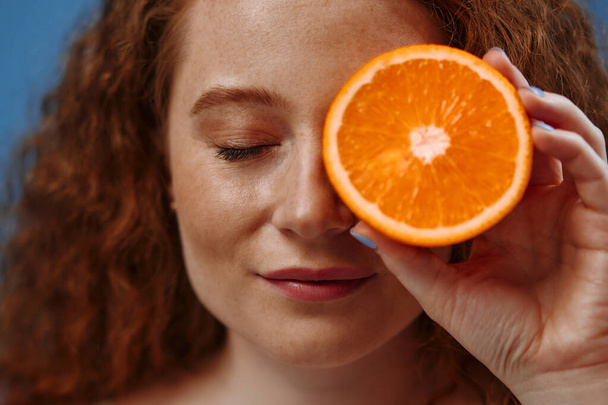 Freckled redhead plump plus size woman in white lingerie holding half an orange isolated on blue background. The concept of face and body skin care, body positive and cosmetology. - Photo, Image