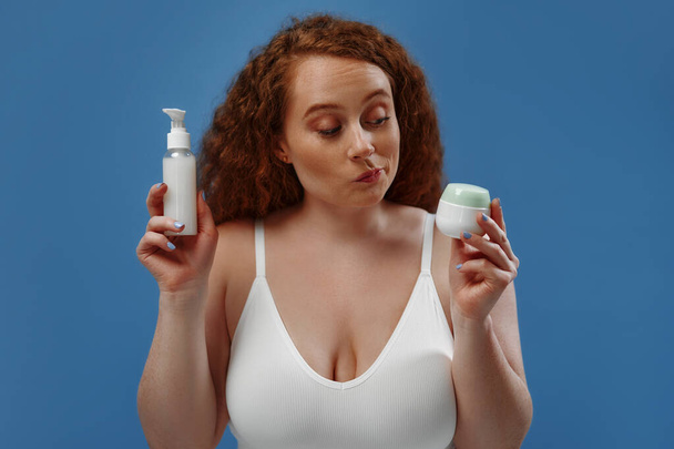 Freckled redhead plump plus size woman in white lingerie holding two jars of cream, not knowing what to choose isolated on blue background. The concept of skin care, body positive and cosmetology. - Фото, зображення