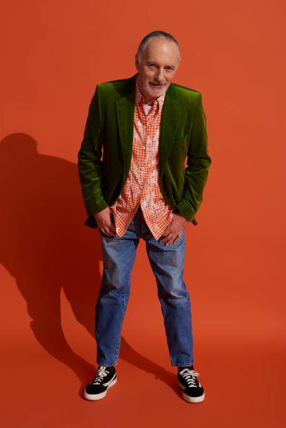 homme âgé positif en chemise et veste en velours vert tenant les pouces dans des poches de jeans bleus, penché vers l'avant et regardant la caméra sur fond rouge orange, concept de vieillissement élégant - Photo, image