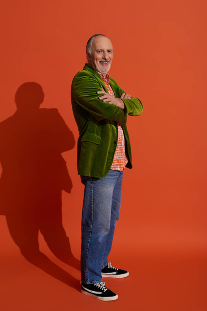 volledige lengte van de knappe en bebaarde senior man met gevouwen armen en lachend naar de camera op rood oranje achtergrond met schaduw, groene velours blazer, blauwe denim jeans, stijlvol verouderingsconcept - Foto, afbeelding