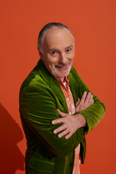 optimistic and charismatic bearded senior man standing with folded arms and smiling at camera on red orange background, fashion look, personal style, green velour blazer, happy aging concept - Photo, Image