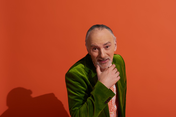 modny i charyzmatyczny starszy mężczyzna w zielonym welurowym blezerze dotykający brody i uśmiechający się do kamery podczas pozowania na czerwonym tle, radosne emocje, pozytywna i modna koncepcja starzenia się - Zdjęcie, obraz
