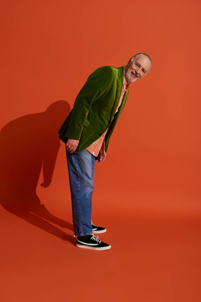 full length of goyful and bearded senior man in trendy casual clothing standing and looking at camera on red orange background, πράσινο βελούδινο σακάκι, μπλε τζιν τζιν, χαρούμενη αντίληψη γήρανσης - Φωτογραφία, εικόνα