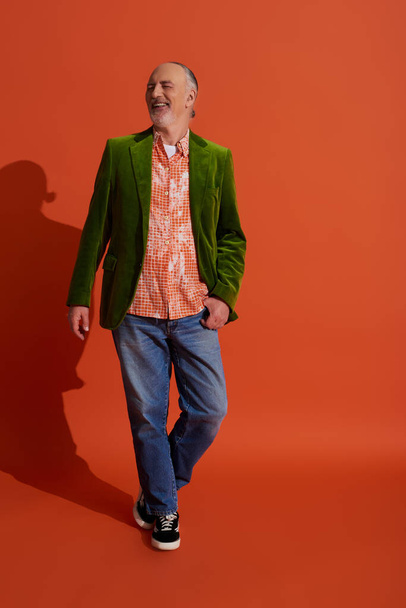 piena lunghezza di uomo anziano positivo in camicia alla moda e blazer di velluto verde in posa con pollice in tasca di jeans denim blu mentre in piedi su sfondo arancione rosso, elegante concetto di invecchiamento - Foto, immagini
