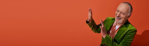 happy and trendy aging concept, excited grey haired and bearded senior man in green velour blazer showing stop gesture and laughing with closed eyes on red orange background, banner with copy space - Photo, Image