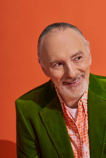 stylish aging and positive lifestyle concept, portrait of optimistic senior male model smiling and looking away on vibrant orange background, grey hair, groomed beard, green velour blazer - 写真・画像