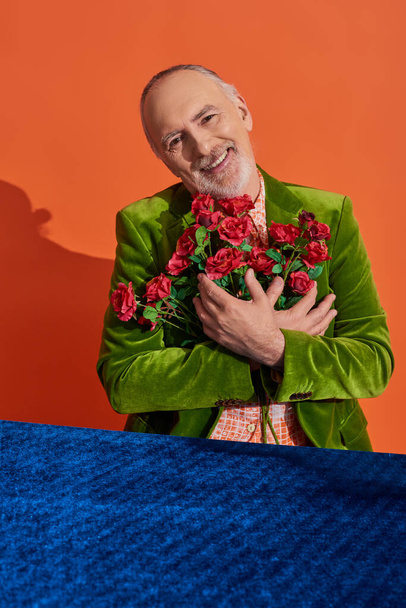 modelo masculino senior lleno de alegría en chaqueta de terciopelo verde abrazando ramo de rosas rojas y sonriendo a la cámara cerca de la mesa con tela de terciopelo azul sobre fondo naranja vibrante, concepto de envejecimiento feliz - Foto, Imagen