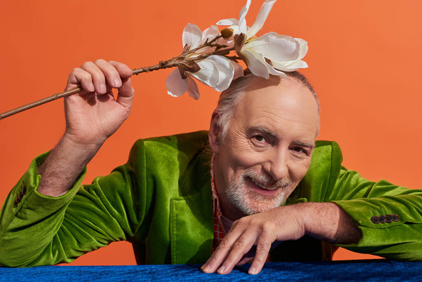 happy aging concept, opgetogen en bebaarde senior man in groen fluwelen blazer zittend aan tafel met blauwe fluwelen doek en met witte orchidee bloem boven het hoofd op levendige oranje achtergrond - Foto, afbeelding