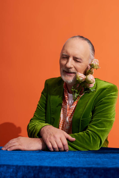ευχαριστημένος ηλικιωμένος άνδρας χαμογελώντας με κλειστά μάτια, ενώ κάθεται με φρέσκα τριαντάφυλλα κοντά στο τραπέζι με μπλε βελούδινο πανί σε ζωντανό πορτοκαλί φόντο, πράσινο βελούδινο σακάκι, στυλ πρόσωπο, χαρούμενη αντίληψη γήρανσης - Φωτογραφία, εικόνα