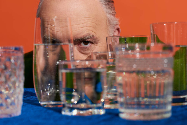 Senior männliches Modell mit ausdrucksstarkem Blick in die Kamera hinter verschwommenen Kristallgläsern mit reinem Wasser auf orangefarbenem Hintergrund, alternde Bevölkerung, Symbolik, Lebensfüllungskonzept - Foto, Bild