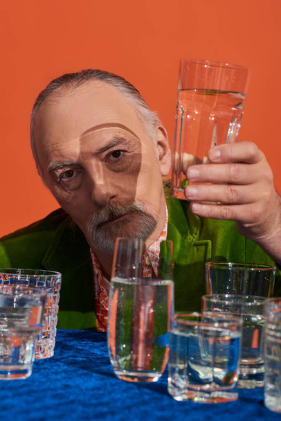 senior bearded man in fashionable and green velvet blazer holding glass of clear water and looking at camera on orange background, aging population, symbolism, life fullness concept - Photo, Image