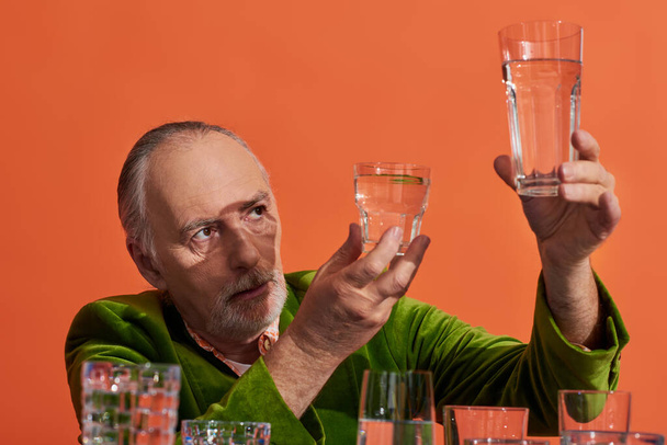 nachdenklicher Mann mit grauen Haaren und Bart, grüner Samtblazer, Brille mit reinem Wasser auf orangefarbenem Hintergrund, alternde Bevölkerung, Symbolik, Lebensfüllungskonzept  - Foto, Bild
