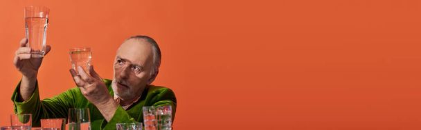 stylish senior man in green velvet blazer looking at glasses with clear water on orange background, aging population, symbolism, life fullness concept, banner with copy space - Photo, Image