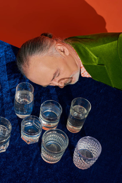 high angle view or aged grey haired man with closed eyes laying on table with blue velour cloth and glasses with pure water on orange background, aging population, symbolism, life fullness concept - Photo, Image