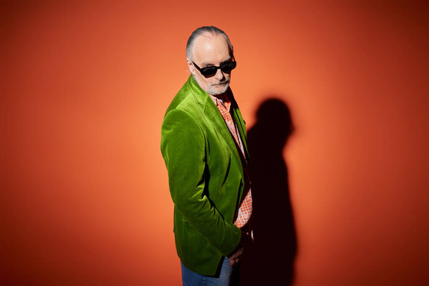 senior male model in stylish casual clothes looking at camera on red and orange background with shadow, green velour blazer, dark sunglasses, expressive individuality, fashionable aging concept - Photo, Image
