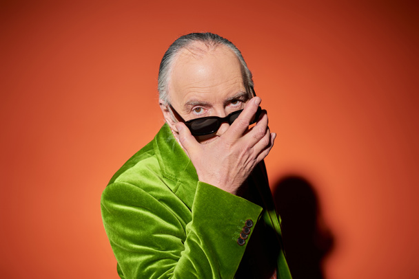 elderly and fashionable man in green velour blazer taking off dark stylish sunglasses and looking at camera with expressive gaze on red and orange background, trendy aging concept - Photo, Image