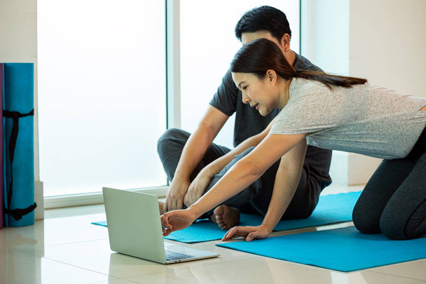 Two Asian couple husband wife sitting on blue mat looking VDO from laptop practice Yoga pose in gym - Фото, зображення
