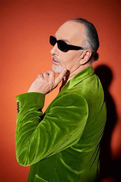 pensive senior man with grey hair and beard, in stylish casual attire touching face and looking away on red and orange background with shadow, dark sunglasses, green velour blazer, fashion and age - 写真・画像