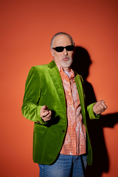 cool senior man in dark sunglasses, trendy shirt and green velour blazer gesturing and pouting lips while having fun and posing on red and orange background with shadow, fashionable and happy aging - Fotó, kép
