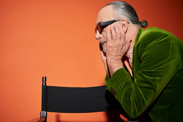 side view of senior grey haired model touching face and looking away near chair on red and orange background, fashion look, dark sunglasses, green velour blazer, fashionable aging concept - Photo, Image